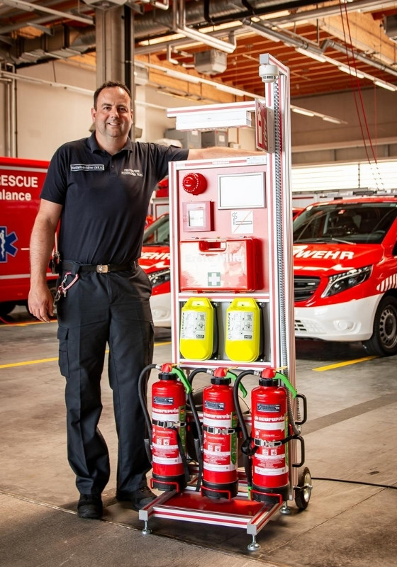 Feuerwehr Rollcontainer Erste Hilfe vor Löschfahrzeug – mit Feuerwehrmann Ein Feuerwehrmann steht neben einem Feuerwehr Rollcontainer für Erste Hilfe vor einem Löschfahrzeug auf dem Gelände der Feuerwehrwache.
